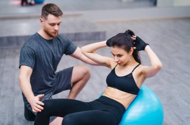 Çekici genç kadın fitness Ball ABS spor salonunda yerde kişisel antrenör ile yapıyor. Sportif kız başını ve güçlü bir erkek Koç arkasında silah ile bir top üzerinde oturan vücudu eğilir