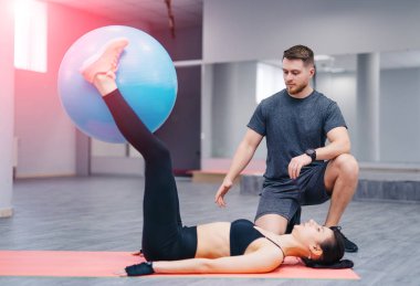 Mindere yalan ve mavi fitness topu bacaklar ile spor salonunda tutarak sağlıklı genç sporcumuz. Kas eğitmen bir spor kulübü olarak istemcinin çalışma kontrol eder.