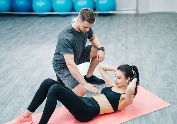 Sporty woman doing abs on the floor and a male trainer holding hand over her stomach in the gym. Handsome instuctor helps pretty female during the training