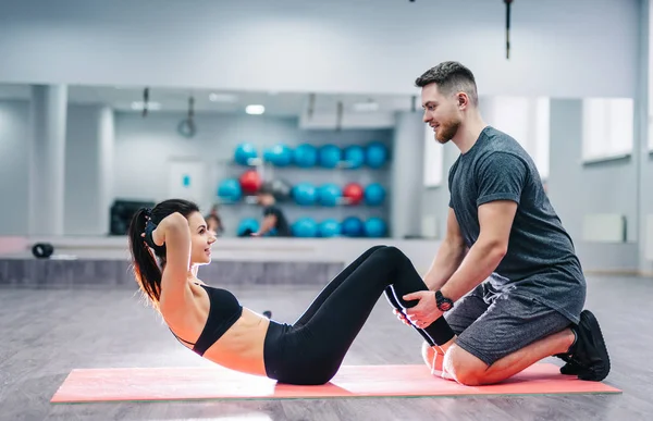 Side view of a girl doing press exercises on the mat with the help of a smiling male. Happy man and woman in a good sport shape doing exercises.
