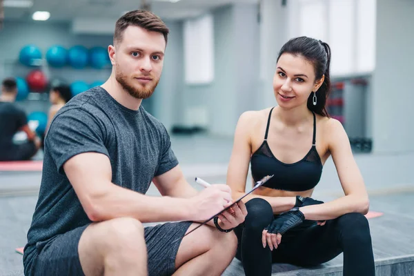 Portrait of a handsome man and attractive woman resting after workout and looking into camera in sports gym. Personal instructor with a client making a training plan