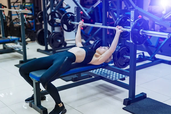 Professional sporty woman builds muscle arms and chest on the simulator in the gym. Athletic girl with a perfect body lifting metal barbell. Concept of a healthy lifestyle.
