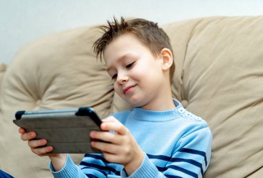 Yumuşak bir koltukta oturan ve evde modern bir aygıt ilginç video izlerken mutlu çocuk. Mavi kazaklı bir dijital tablet kullanarak ifade güler yüz, küçük çocuk