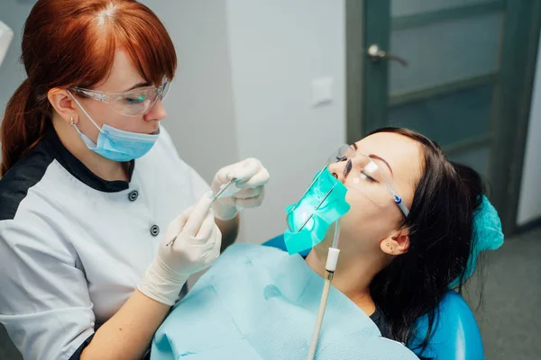 Mujer dentista en guantes médicos trabajando en los dientes de sus pacientes en la sala de cirugía dental. Mujer joven acostada con los ojos cerrados en una silla de dentista y el especialista la trata . —  Fotos de Stock