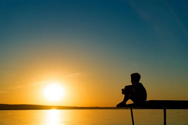 Niño sentado solo en la pasarela sosteniendo sus piernas y soñando al atardecer. Pequeño niño observa la puesta de sol sobre el río . — Foto de Stock