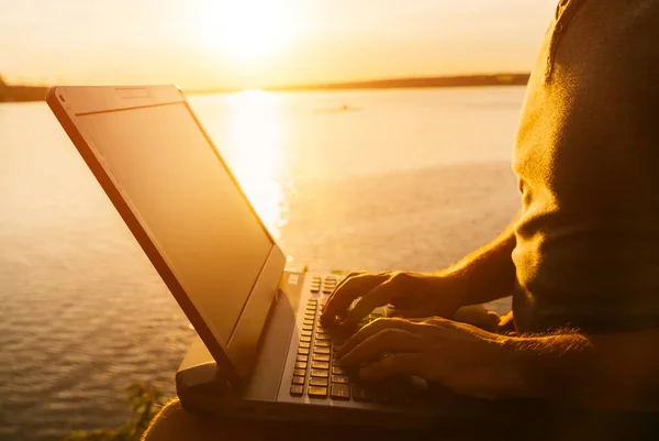 Mann arbeitet abends an seinem Laptop im Freien am Flusshintergrund. Der Mann tippt bei Sonnenuntergang mit den Händen auf einem Laptop. Nahaufnahme — Stockfoto