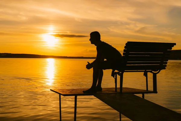 Silhouette of a man sitting on the bench in a bridge and looking at beautiful orange sunset. Single man watching sunset over a lake and thinking about his life in summer. — Stock Photo, Image