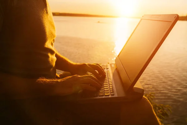 Mannens händer arbetar på en bärbar dator på kvällen nära floden vid solnedgången. Arbeta under semester utomhus på sommaren. Närbild — Stockfoto