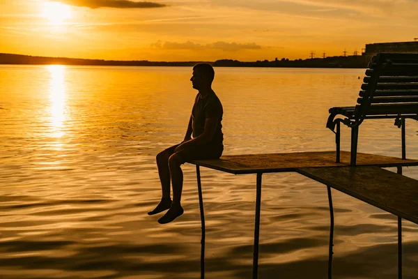 Ein verträumter Mann sitzt am Rande eines Mauerwerks über dem Wasser bei einem wunderbaren Sonnenuntergang. fantastische Aussicht auf einen Sonnenuntergang mit einem einzigen Mann, der sich in der Nähe des ruhigen Flusses entspannt. — Stockfoto