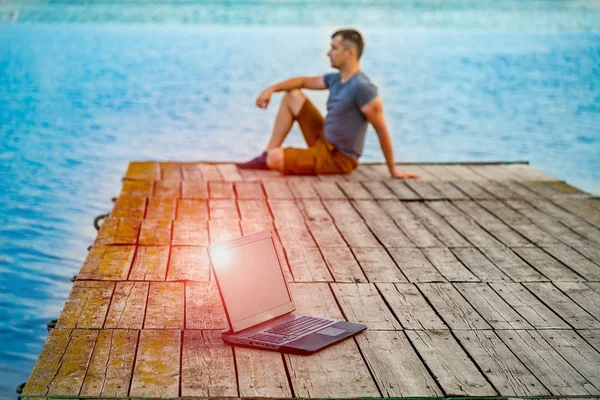 Work and travel. Young man using laptop computer sitting on wooden fishing pier with beautiful sea view.