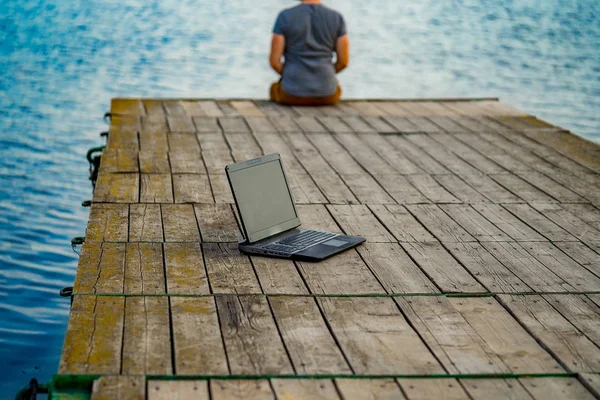 Ung man sitter på en brygga, med sin laptop. Arbete och semester. — Stockfoto