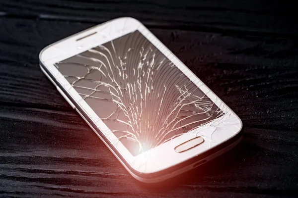 Damaged screen of a broken smartphone isolated on dark background. Crashed mobile phone lying on the floor inside. Close-up