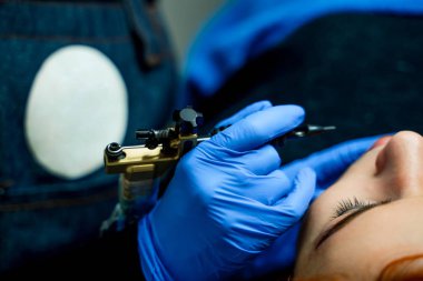 Kalıcı makyaj mavi eldiven profesyonel güzellik uzmanı elinde siyah aleti. Genç kadın kalıcı dudak makyaj oluyor. Yakın çekim