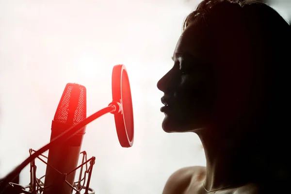 Perfil de silueta sensual joven con cara bonita cantando al micrófono retro sobre fondo blanco. Mujer sexy delante del micrófono con fugas de luz roja en el estudio. Primer plano —  Fotos de Stock