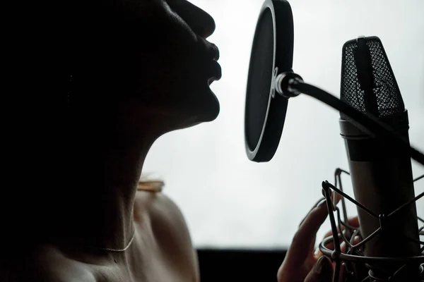 黑暗的剪影外形一个年轻的女性与赤裸的肩膀唱歌在被隔绝的话筒在演播室。面对一个性感的女人表演音乐靠近麦克风。特写 — 图库照片
