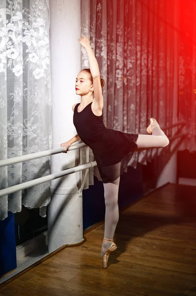 Hermosa bailarina joven en traje negro está de pie en pose en sus puntillas en el estudio. Chica bonita sostiene su pierna y brazo cerca del soporte en el suelo de madera . —  Fotos de Stock