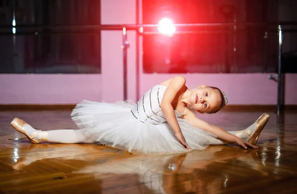 Beautiful preschool girl in a classical ballet tutu stretching her body on the wooden studio floor. Pretty little ballerina doing exercises on the floor and looking at camera. — Stock Photo, Image