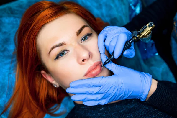 Professionele tatoeëerder maken van permanente make-up op lippen voor mooie roodharige vrouw. Mooie jonge vrouw die het verkrijgen van de tatoeage op gezicht in salon. — Stockfoto