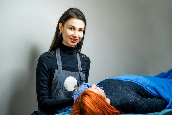 Beau cosmétologue regardant la caméra près du patient dans un salon de beauté. Cosmétique appliquant un maquillage permanent sur les lèvres de la femme sur un fond mural gris . — Photo