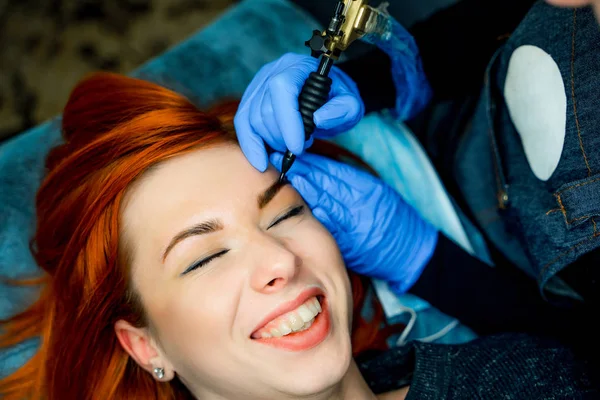 Woman lying on couch at beauty salon with closed eyes and grinning. Professional doctor wearing blue gloves and holding speciality tool making eyebrows permanent make-up. Close-up — Stock Photo, Image
