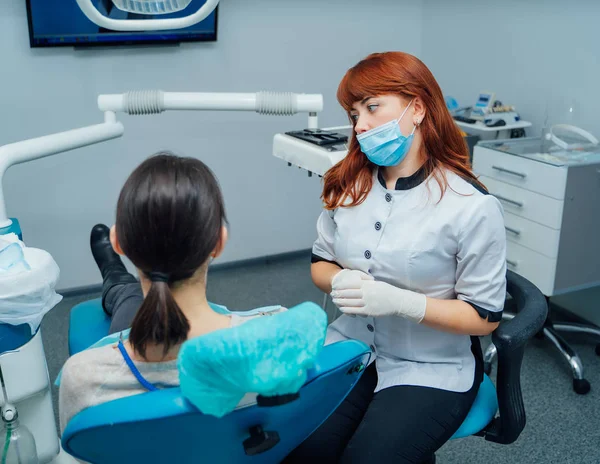 Estomatologista ruivo bonito na máscara que fala ao paciente no contexto de clínica moderno. Jovem dentista e uma paciente falando . — Fotografia de Stock