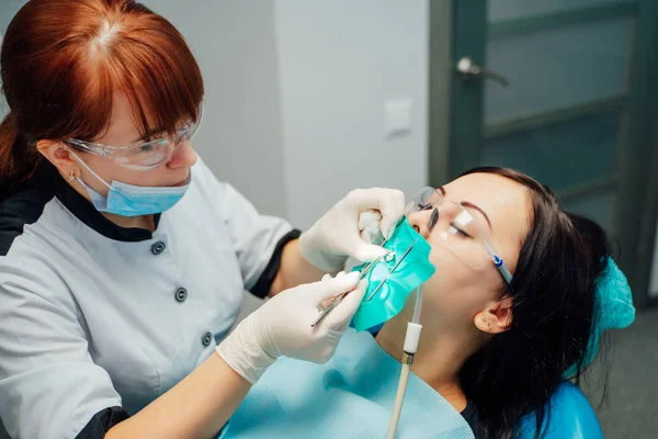 Zbliżenie dentysta Kobieta w bieli, uniform, ochronne okulary o pracy w kliniki stomatologiczne. Traktuje pacjenta kobieta z zamkniętymi oczami siedząc w krzesło lekarskie i dentysta zęby — Zdjęcie stockowe