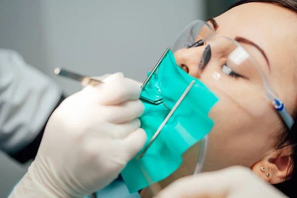 Paciente feminina em óculos de proteção com olhos fechados em tratamento em consultório odontológico. Estomatologista profissional em luvas estéreis está trabalhando com o paciente na clínica. Close-up — Fotografia de Stock