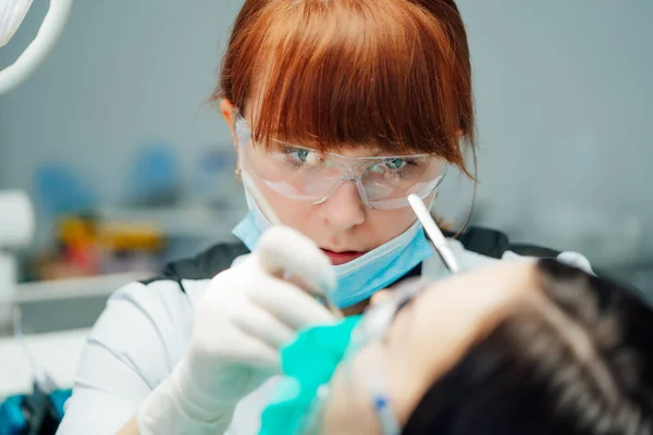 Rothaarige Zahnärztin mit Schutzbrille und Maske, die die Zähne des Patienten in der Klinik untersucht. Zahnarzt Kariesbehandlung in der Zahnarztpraxis. Nahaufnahme — Stockfoto