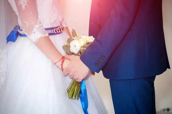 Hermosa pareja joven de pie uno junto al otro durante la ceremonia de la boda. Novia en vestido blanco con cinturón azul y novio llevando un elegante traje tomados de la mano . — Foto de Stock