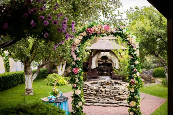 Prachtige boog versierd met kleurrijke bloemen op de achtergrond van een kleine fontein buitenshuis. Natuurlijke achtergrond in het park met bloemen boog, groen gras en bomen in een zonnige dag. — Stockfoto