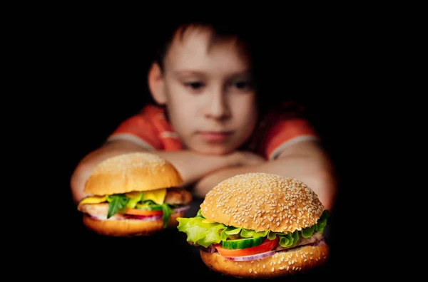 Милый мальчик сидит перед гамбургерами и думает съесть их на черном фоне. Маленький ребенок и вкусная фаст-фуд. Крупный план — стоковое фото
