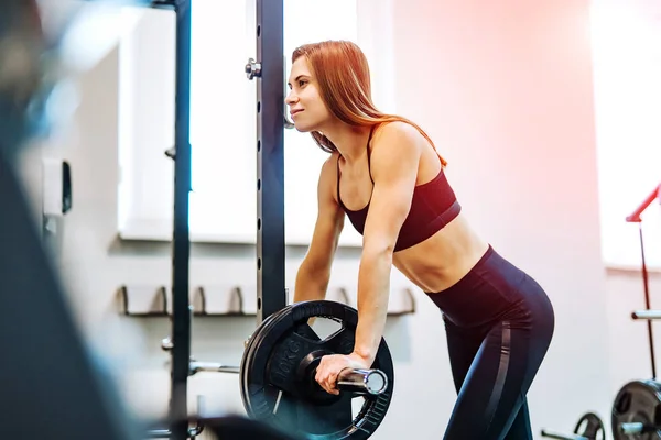 美丽快乐的年轻女运动员穿着健身服站在健身房沉重的杠铃附近。微笑的女性做举重锻炼在体育中心背景. — 图库照片