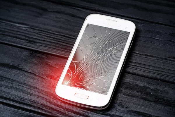 Modern smartphone with large broken screen with debris. Damaged expensive device on the dark wooden surface with light leaks. Close-up