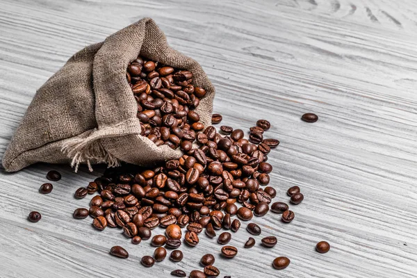 Bag of natural brown coffee beans and some scattered seeds on the light wooden background. Roasted coffee grains spilling out of sack on the table.