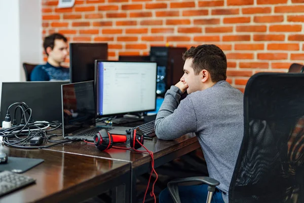 Serious young programmer sitting at his worktable inside the office and facing a computer monitor. Creative worker holds his hand on his mouth and concentrates on problem.