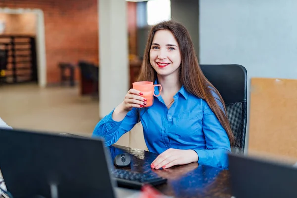 Young woman programmer sitting in a company office in front of the computer with a pink cup. Professional computer engineer looking at camera and smiling. Software work