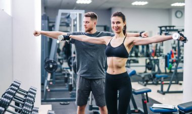 Siyah üst ve tozluk içinde gülümseyen kadın spor salonunda antrenörü önünde dumbbells tutarak. Erkek eğitmen ve güzel kadın metal halter kaldırma ve kamera bakıyor.