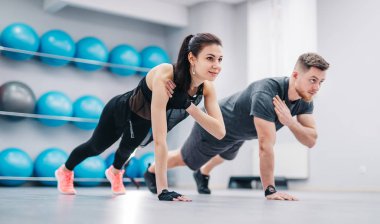 Genç bir çift, spor toplarının arka planında yerden şınav çekiyor. Spor salonunda spor giyinen erkek ve kadın sporcular..