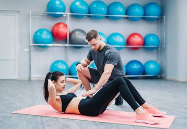 Spor salonundaki minderde koçunun yardımıyla karın egzersizi yapan genç bir kadın. Kas antrenörü çekici bir kadının fitness toplarının arka planında karın kası yapmasına yardım eder..