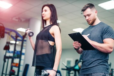 Genç kadın spor salonunda dambıllarla egzersiz yaparken panoya erkek yazı. Kişisel antrenör spor merkezinde ince bir kadın sonuçlarını kontrol eder.