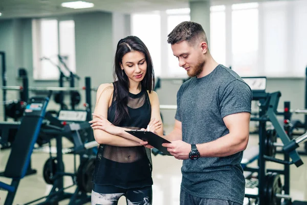 Muskeltrainer zeigt die Sportergebnisse der Frauen vor dem Hintergrund des Fitnessstudios. Personal Trainer und junge Frauen im Gespräch über die Fortschritte im Sportverein. — Stockfoto