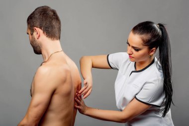 Gri arka plandaki bir adamın arkasını inceleyen güzel hemşire. Beyaz tıbbi ceketli esmer kadın doktor hastanın sırtını kontrol ediyor.