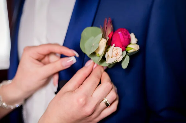 Las manos de la novia sujetando un pequeño boutonniere a una chaqueta para el novio. Decoración floral tradicional para novio, un accesorio en traje de boda festivo. Primer plano — Foto de Stock