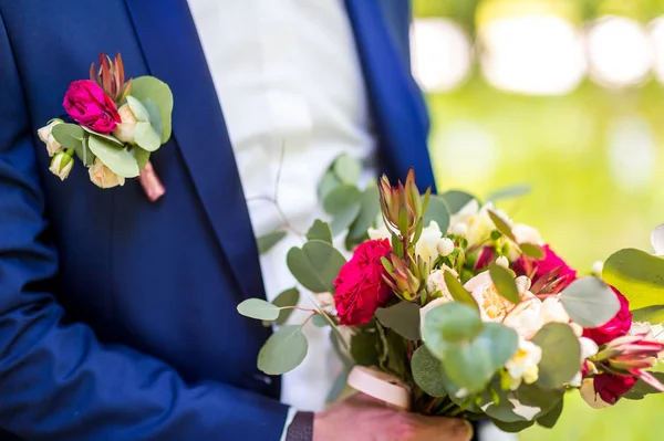 Beautiful bouquet of mixed flowers in the hands of a groom in blue suit. Wedding bunch of fresh flowers for bride and a small cute boutonniere pinned to a wedding suit on groom. — Stock Photo, Image