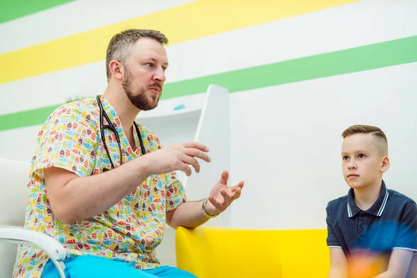 Un bel dottore maschio cura un ragazzo in clinica. Bambino piccolo e pediatra professionista in abiti medici sono seduti nella stanza. Sanità e medicina . — Foto Stock