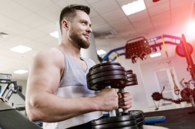 Yakışıklı gülümseyen adam crossfit spor salonunda iki ağır dumbbells ile oturuyor. Güçlü vücut geliştirmeci simülatörleri ile spor kulübünün arka planda dumbbells tutuyor.