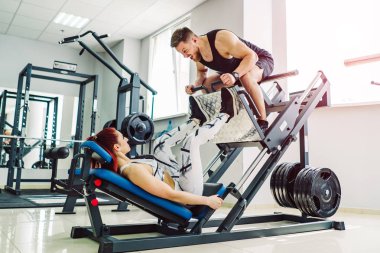 Genç bir kadın modern bir simülatörle meşgul ve bir adam spor salonunda oturuyor. Kız bir antrenörle antrenman yapıyor. Bayan sporcu, antrenman makinesiyle bacak egzersizleri yapıyor..