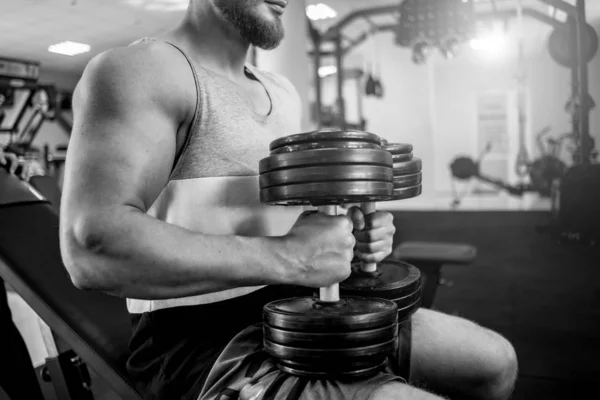 Güçlü adam vücut geliştirmeci spor salonu arka plan üzerinde ağır dumbbells ile oturuyor. Spor merkezinde bir sporcunun kaslı vücut. Siyah beyaz fotoğraf. — Stok fotoğraf