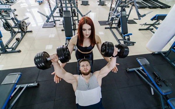 Güçlü bir adam halter ve güzel bir kadın kaldırarak üst görünümü spor salonunda ona yardımcı olur. Aktif çift modern bir spor kulübünün arka planda dumbbells ile egzersiz. — Stok fotoğraf