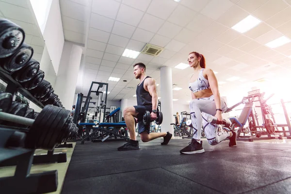 Pareja sana en ropa deportiva levantando pesas en el gimnasio. Mujer atractiva y hombre guapo haciendo ejercicio con pesas de pie en pose especial en el club deportivo. — Foto de Stock